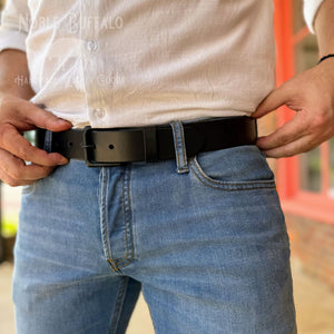 A close-up of a man wearing a black leather belt with a matte black rectangular buckle, paired with light blue jeans and a white button-down shirt. The watermark 'Noble Buffalo' is visible in the upper left.