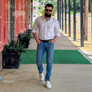 A man wearing a white long-sleeve button-down shirt, blue jeans, a black belt, and white sneakers walks along a covered outdoor corridor next to a brick building with red trim. The image has a 'Jable Studio' watermark overlay.