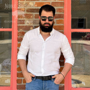 A man with a beard and sunglasses wearing a white long-sleeve button-down shirt and blue jeans, standing in front of a brick wall framed by a red window.