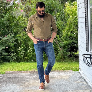 A man wearing a brown button-down shirt, blue jeans, and brown leather shoes walks on a concrete porch with green bushes and a white house in the background.