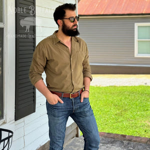 A man with a beard and sunglasses wearing a tan button-down shirt and blue jeans, standing on a porch in front of a house with a green lawn.