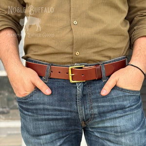 A man wearing a brown leather belt with a gold-tone buckle, tucked into blue jeans, with a khaki button-down shirt. The Noble Buffalo brand logo is visible in the top left corner.