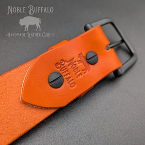 Close-up of the Noble Buffalo maker’s mark and buckle hardware on a carrot orange leather belt
