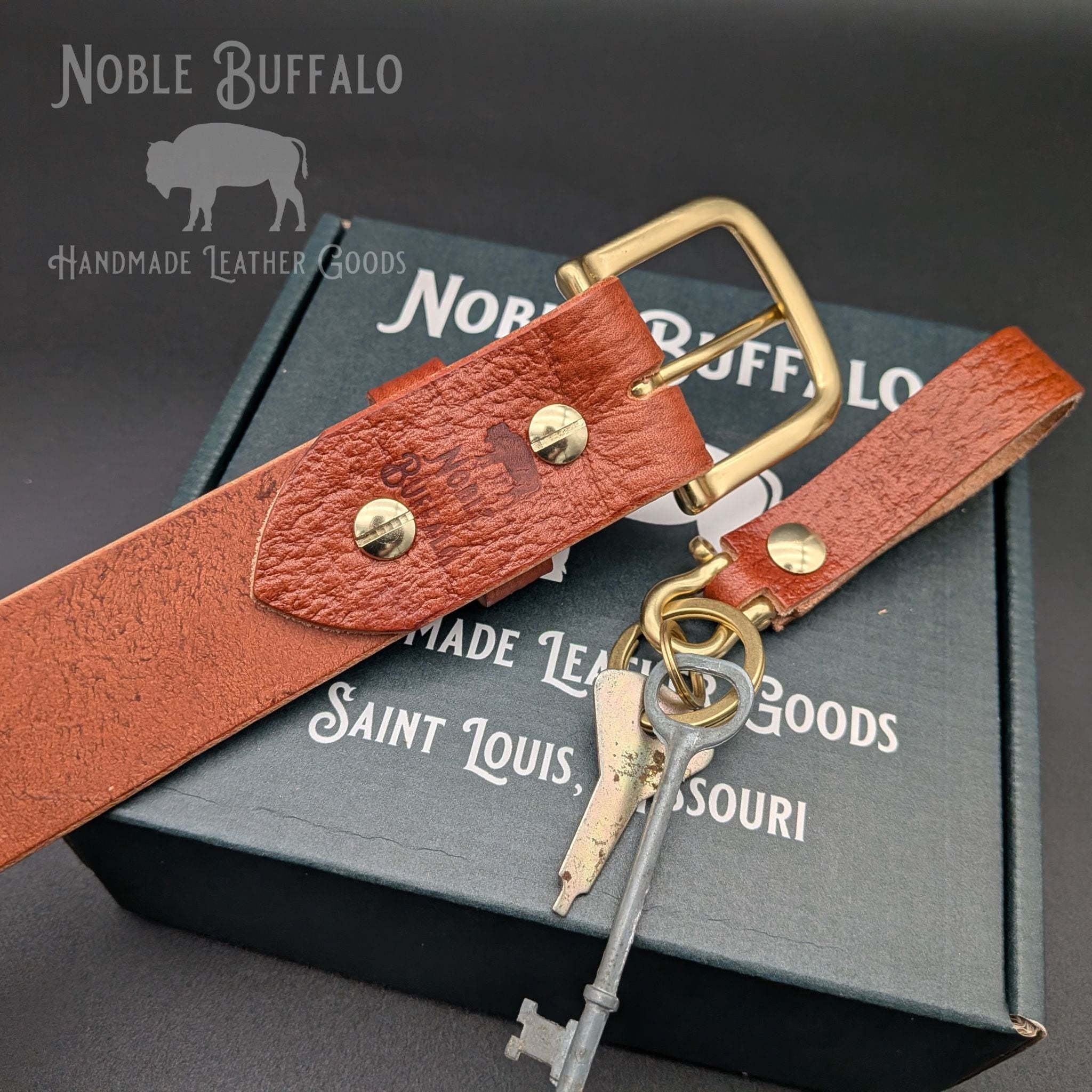 Close-up of chestnut embossed Hermann Oak leather belt with brass buckle and matching keychain on a Noble Buffalo box