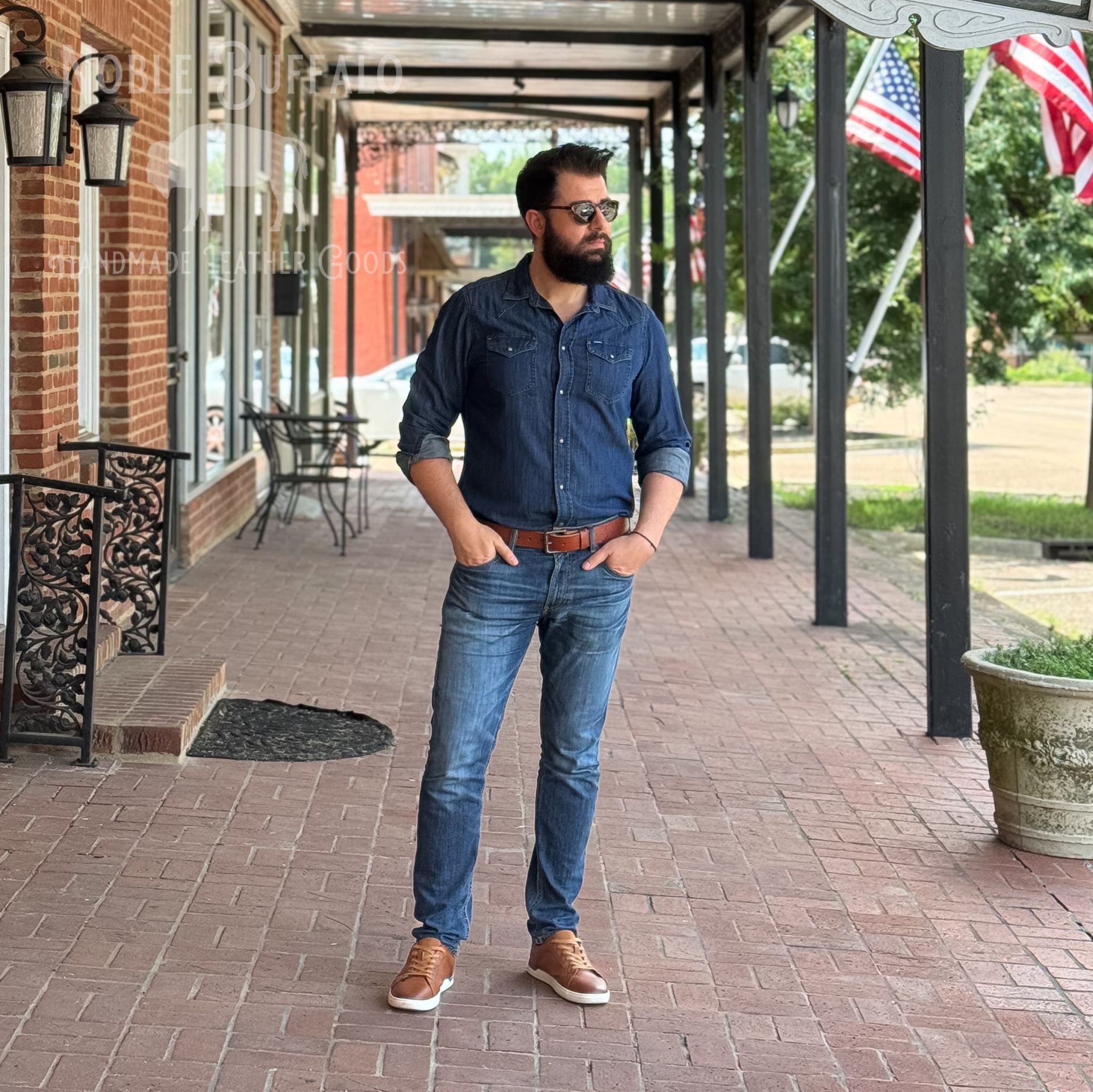 A man wearing a dark blue denim button-up shirt, blue jeans, and brown leather shoes stands on a brick porch walkway.