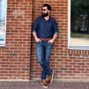 A man wearing a dark blue denim shirt, blue jeans, and brown leather sneakers stands against a red brick wall. The image has a watermark for 'Mobile Buffalo Handmade Leather Goods' on the left side.