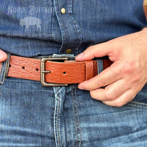 A close-up of a person wearing a tan leather belt with a silver-toned metal buckle, fastened around blue denim jeans. A hand is adjusting the belt.