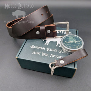 Dark brown Horween Illini Latigo belt displayed on a Noble Buffalo box beside leather balm and a matching leather keychain.