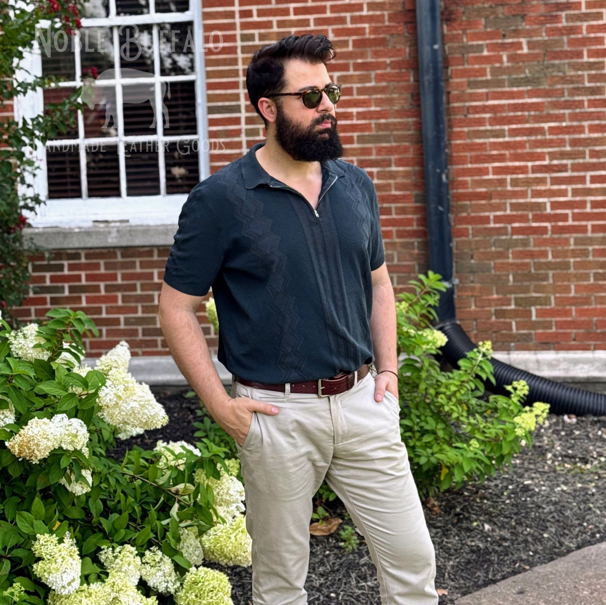 A man with a beard and sunglasses wearing a dark slate blue patterned polo shirt, beige trousers, and a brown belt, standing outdoors in front of a brick building with white flowers.
