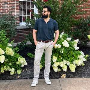 A man wearing a dark slate blue short-sleeve polo shirt, beige trousers, a brown belt, and white sneakers stands outdoors in front of a brick building and green hydrangea bushes.