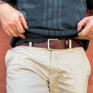 A close-up of a person wearing a dark charcoal knit shirt and beige pants, pulling the shirt slightly outward to reveal a brown leather belt with a silver buckle. The person is standing against a textured terracotta wall.