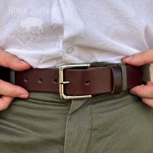 A close-up of a person wearing a dark brown leather belt with a polished gold-tone buckle, paired with olive green trousers and a white shirt. The brand 'Noble Buffalo' is visible as a watermark on the shirt.
