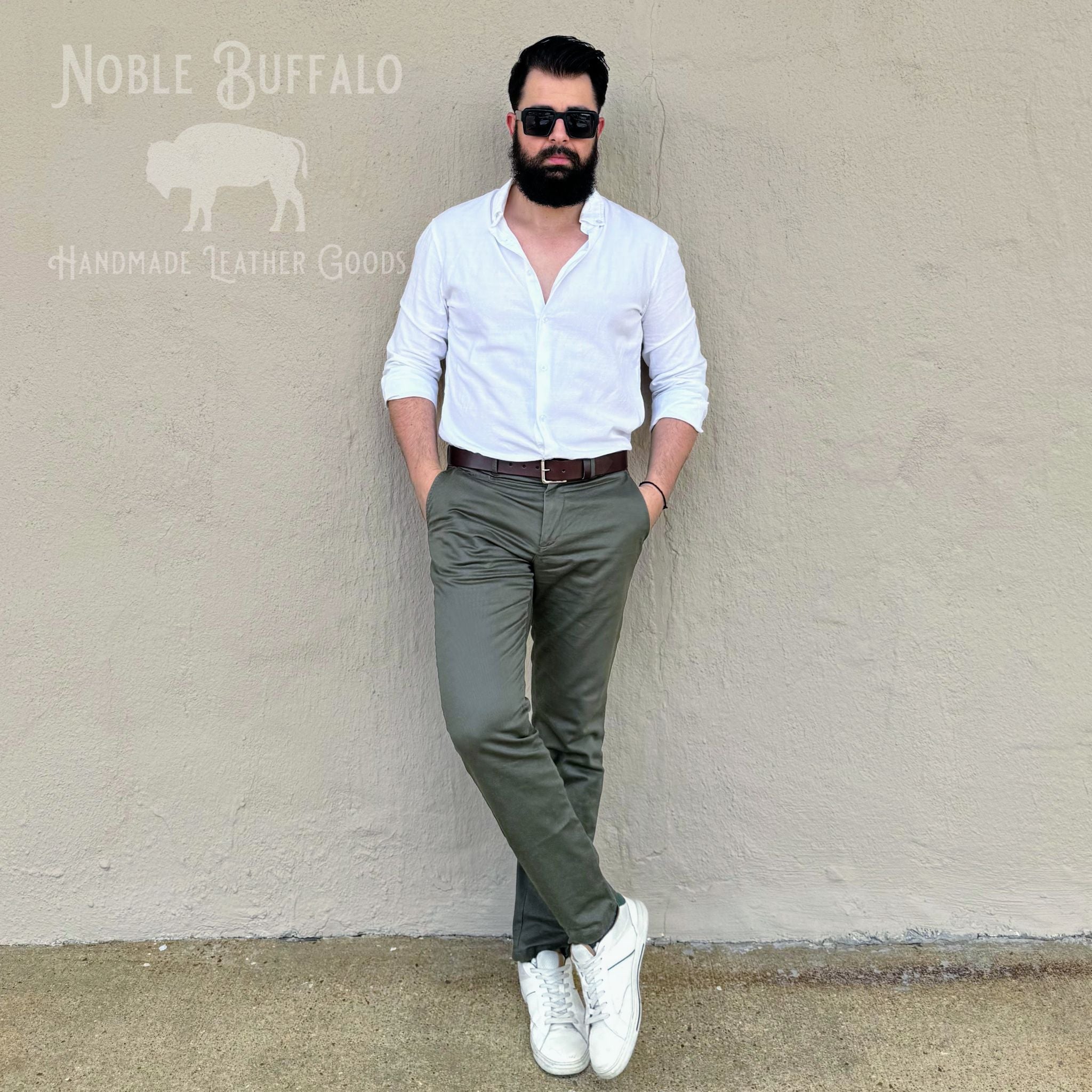A man with a beard and sunglasses stands against a textured beige wall wearing a white button-down shirt, olive green trousers, a brown belt, and white sneakers. A logo for 'Noble Buffalo Handmade Leather Goods' is visible in the top left corner.