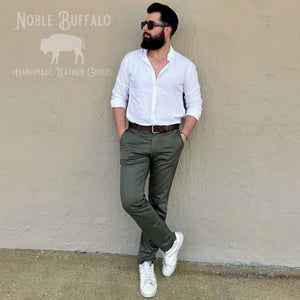 A man wearing a white button-down shirt, olive green trousers, white sneakers, and sunglasses leans against a beige stucco wall. The logo 'Noble Buffalo Handmade Leather Goods' is visible in the top left corner.