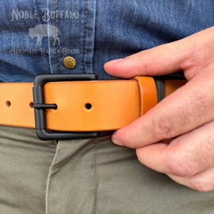 A close-up of a person wearing a tan leather belt with a black metal buckle, fastened at the waist over a blue denim shirt and olive pants. A 'Noble Buffalo Handmade Leather Goods' watermark is visible in the top left.