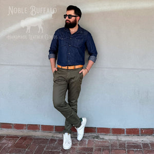 A man wearing a dark blue denim shirt, olive green pants, a tan belt, and white sneakers leans against a gray wall. The image has a 'Noble Buffalo Handmade Leather Goods' logo watermark in the top left corner.