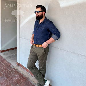 A man with a beard and sunglasses leans against a light gray stucco wall, wearing a dark blue denim button-up shirt, olive green cargo pants, and a tan leather belt. The 'Noble Buffalo Handmade Leather Goods' logo is visible in the top left corner.