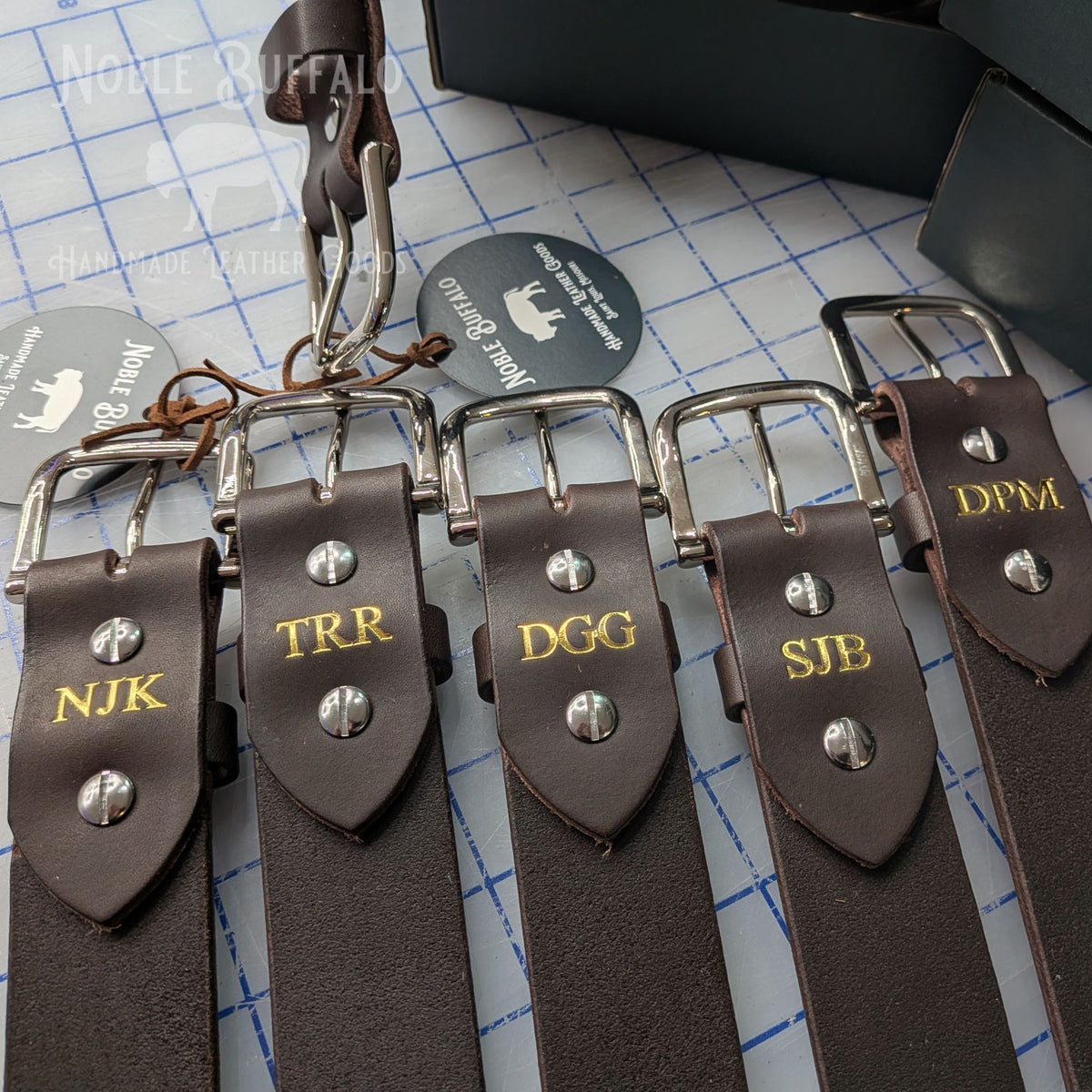 Close-up of leather belt backs embossed with three-letter gold foil initials behind the buckle.