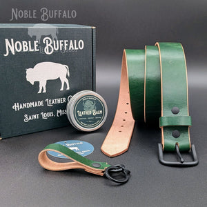 Noble Green Hermann Oak Latigo leather belt with a glossy finish and black buckle, displayed with a matching keychain, leather balm, and Noble Buffalo box.