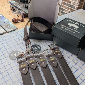 Personalized leather belts with gold foil monograms displayed on a workbench next to Noble Buffalo packaging.