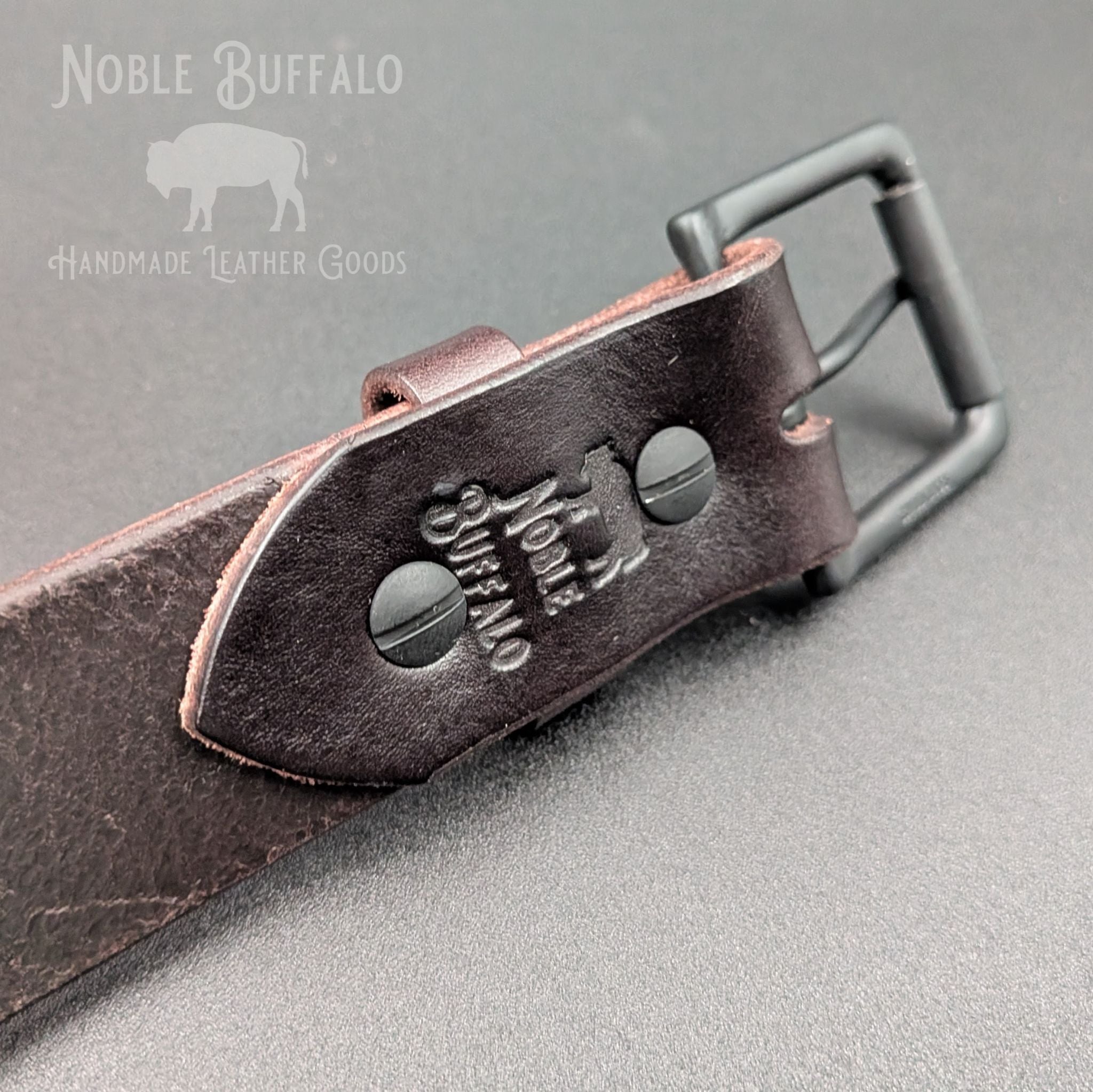Close-up of the Roasted Arabica leather belt showing matte black Chicago screws, buckle, and Noble Buffalo maker’s mark.